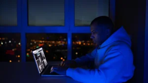 Un uomo lavora al computer portatile di sera accanto a una finestra con le luci della città